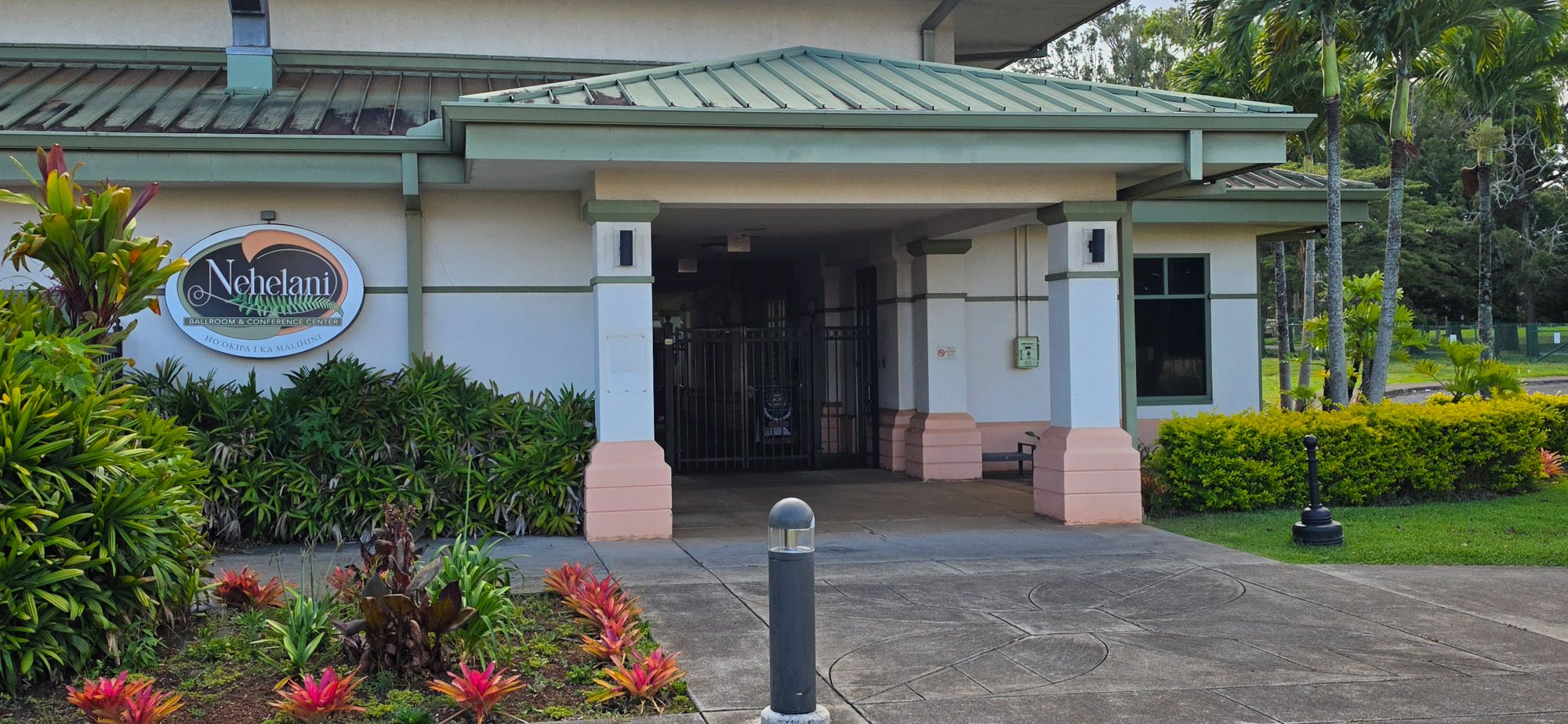 Exterior entrance of the Nehelani Ballroom and Conference Center at Schofield Barracks, Hawaii. The building features a green metal roof, white and pink stucco columns, a covered entryway with iron gate doors, tropical landscaping with colorful plants.